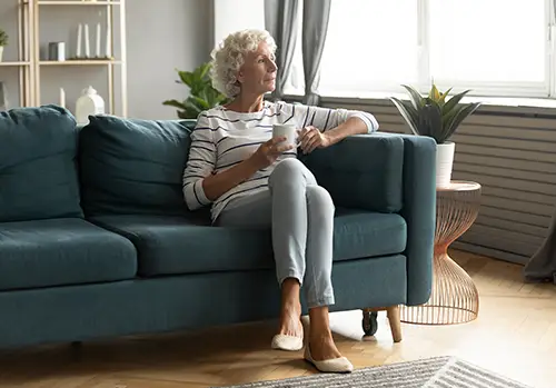 Calm elderly woman sit on comfortable couch in living room drink tea look in window distance thinking or dreaming