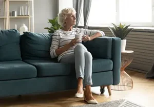Calm elderly woman sit on comfortable couch in living room drink tea look in window distance thinking or dreaming