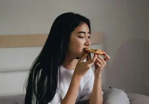 A young woman snacks on pizza in bed.