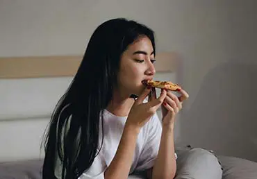 A young woman snacks on pizza in bed.