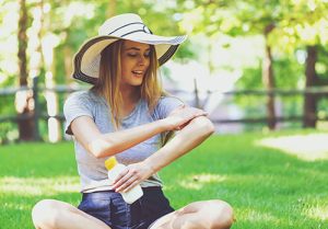 Woman sitting in a sunny park applies sunscreen to her arms.