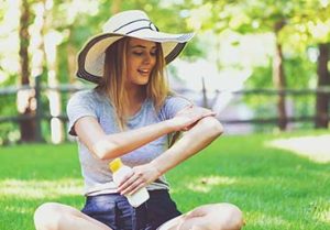 Woman sitting in a sunny park applies sunscreen to her arms.