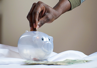 A person's hand places a coin in a nearly empty piggy bank that sits on a bed.