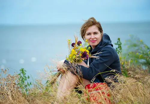 A woman sits and smiles while holding a bouquet of flowers in a field.