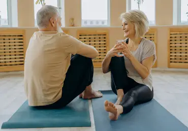 An older couple sit on a pair of exercise mats and smile.