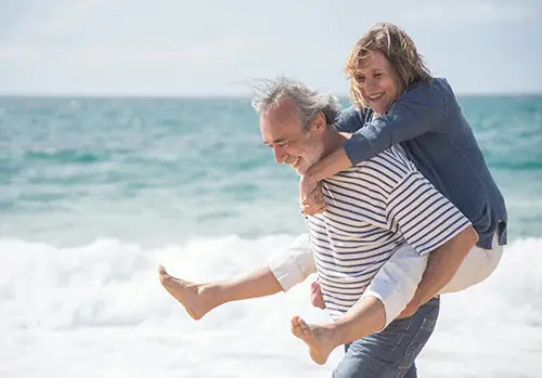 A senior man carries his wife as he walks along a beach.