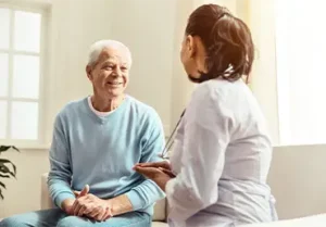 An elderly woman smiling while sitting and holding hands with a female healthcare professional during a conversation in a bright room.
