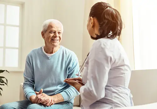 An elderly woman smiling while sitting and holding hands with a female healthcare professional during a conversation in a bright room.