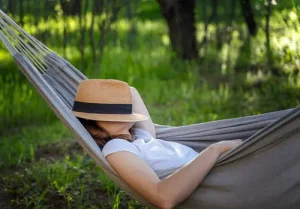 Woman resting in a hammock in a summer garden covering her face with a straw hat.
