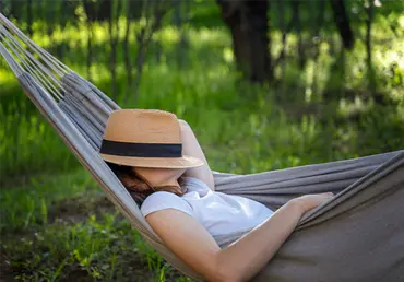 Woman resting in a hammock in a summer garden covering her face with a straw hat.